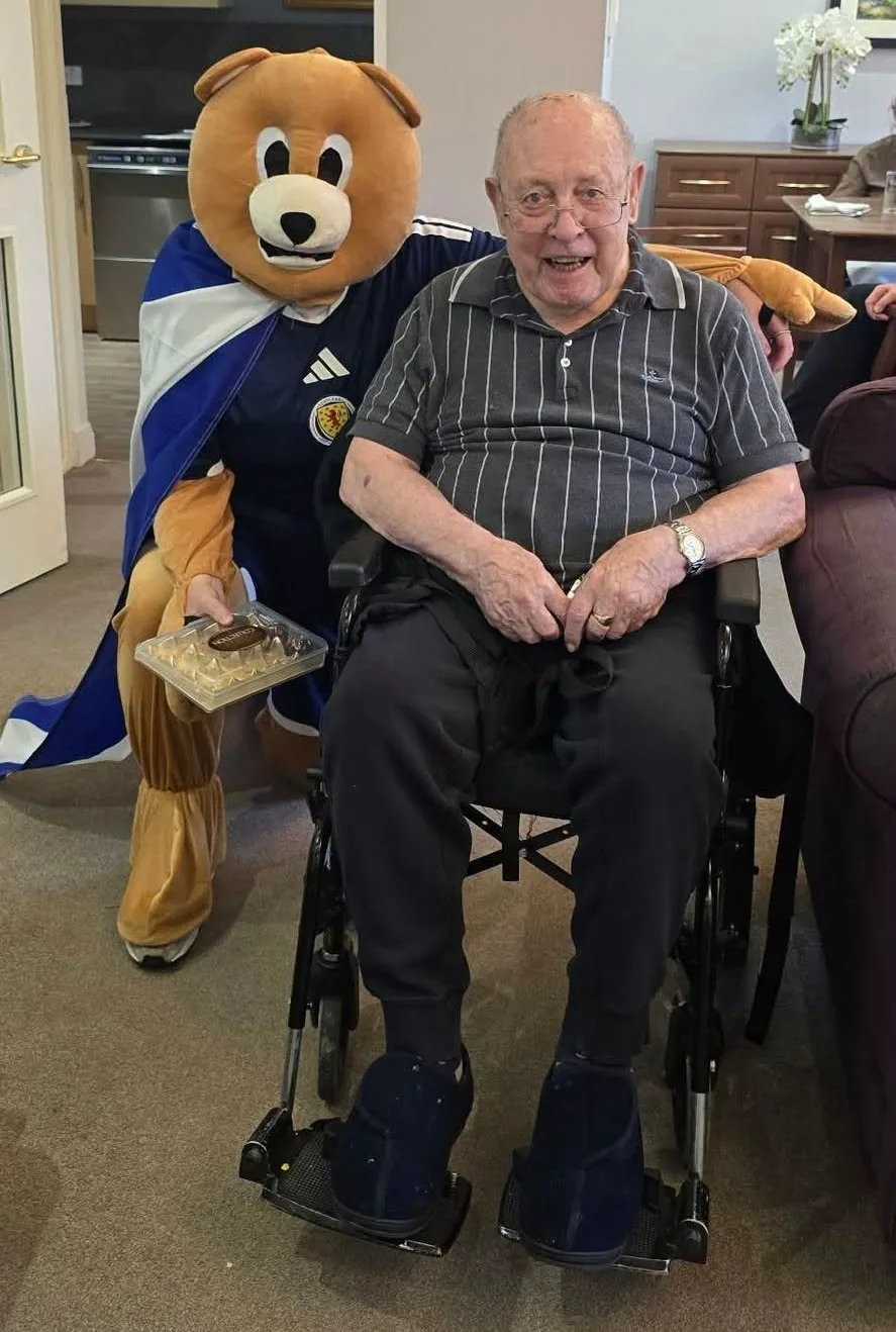 Resident John smiling in his wheelchair beside Roary the mascot, who is holding a box of chocolates, at Culduthel Care Home