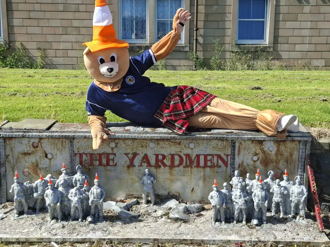 Roary the Big Kick Off mascot wearing a kilt and a traffic cone, sitting behind the Yardmen model display made by residents at Kincaid House Care Home
