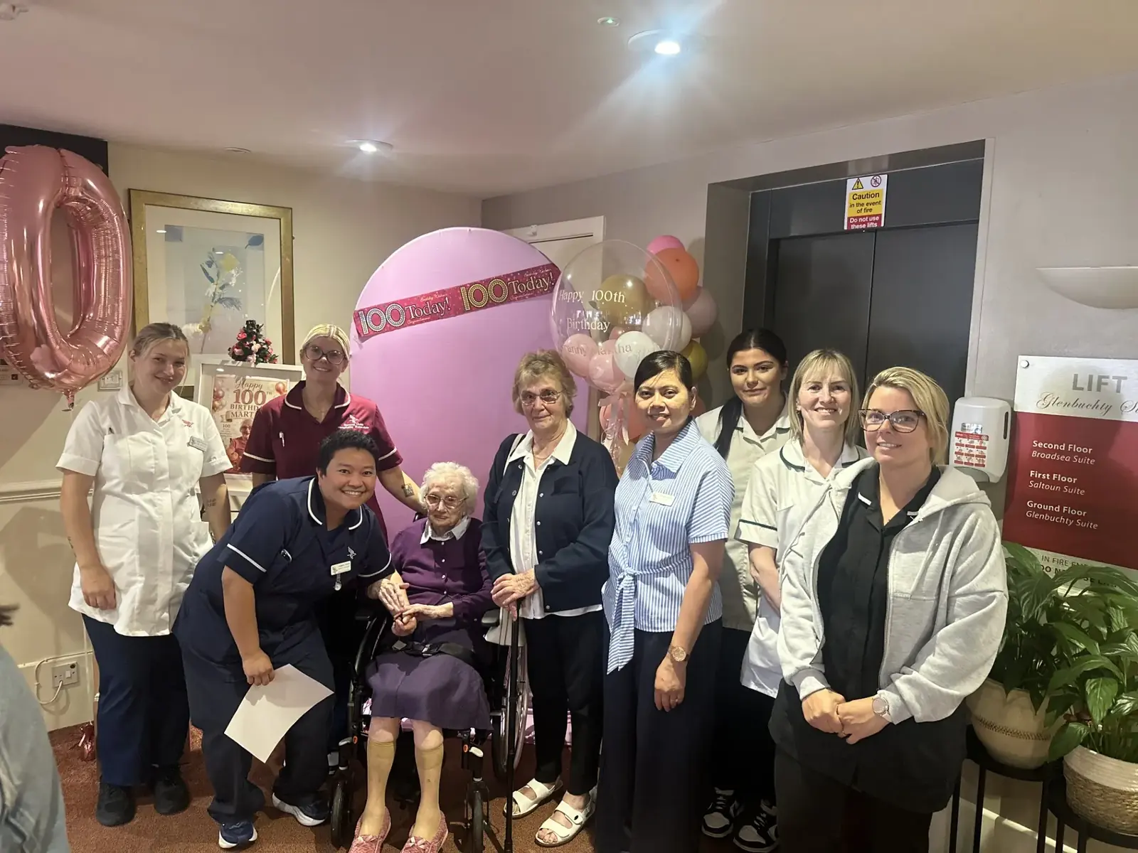 Martha with her family and the Kynnaird House Care Home team beside a 100 Today backdrop and pink balloons