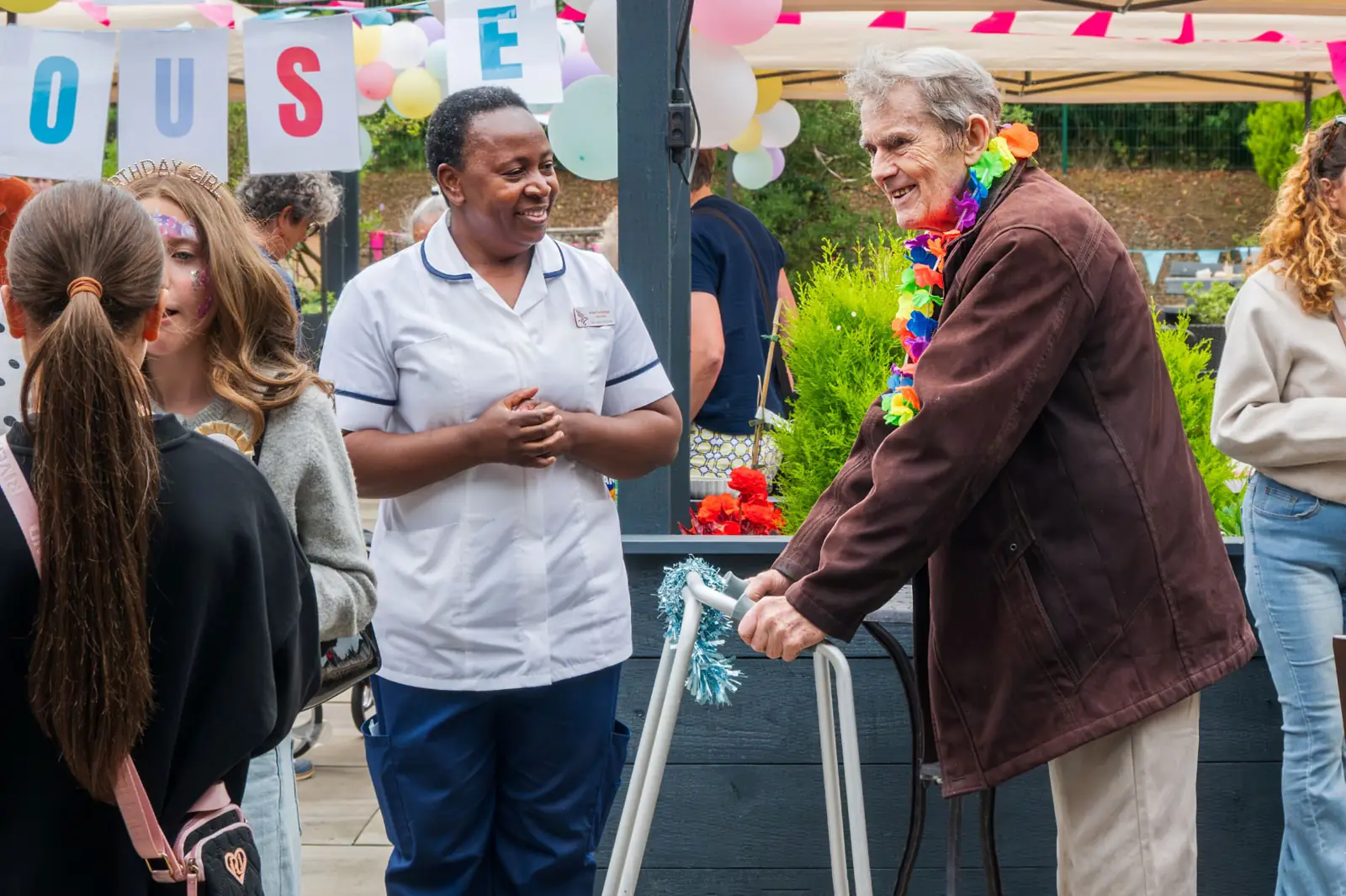 A resident wearing a flower garland talking with a nurse and visitors at the Mearns House summer fete